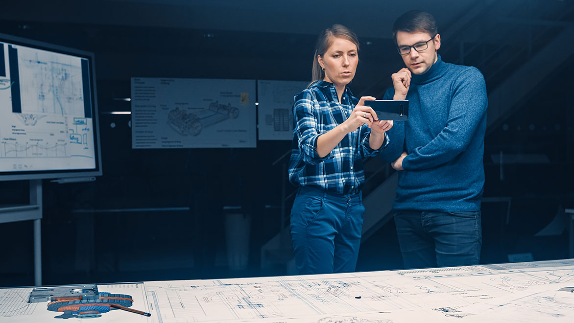 Two young software development engineers look at AR smartphone, with engine blueprints on the desk in front of them 