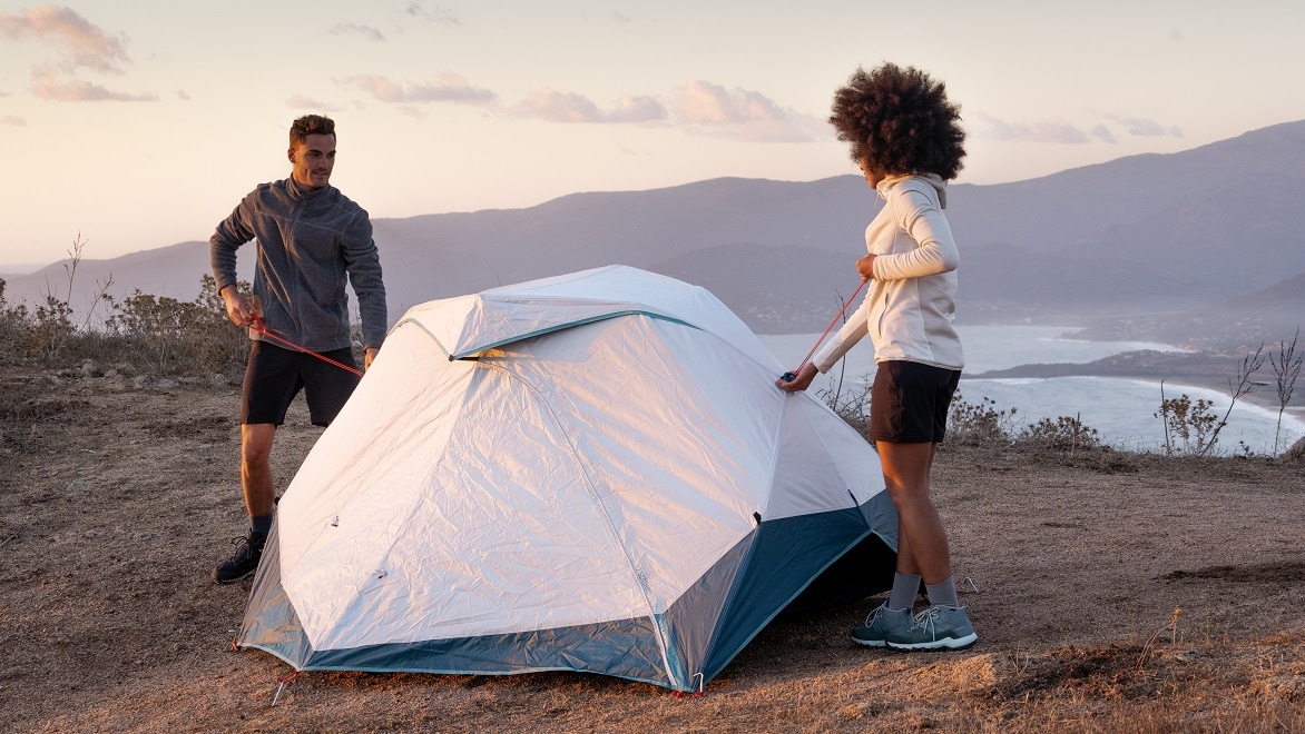 Two people set up a camping tent on a bluff overlooking a lake.