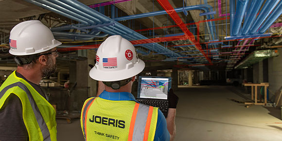  Two people inside a construction site wearing safety gear and using construction software on a tablet 